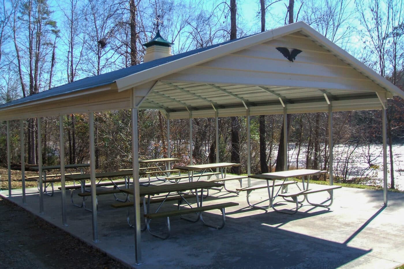 a Open sided carport cover with a gable roof and green cupola, installed on a concrete pad being used to cover picnic tables at a park along the river