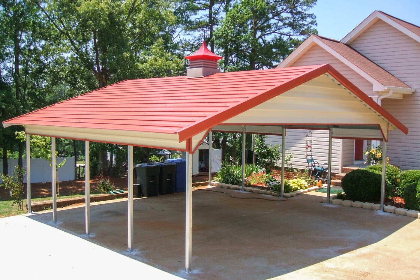A Red roofed carport with a red and gray cupola with cream gabling installed in a concrete driveway next to a house